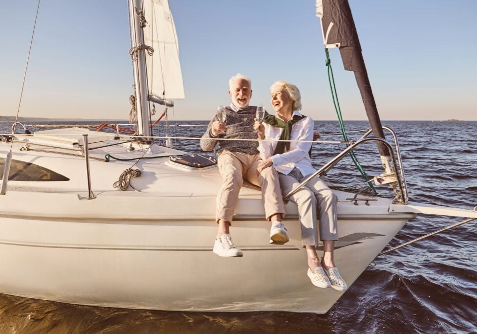 Happy Senior Couple Sitting On The Side Of Sail Boat Or Yacht Deck Floating In Sea. Man And Woman Drinking Wine Or Champagne And Laughing